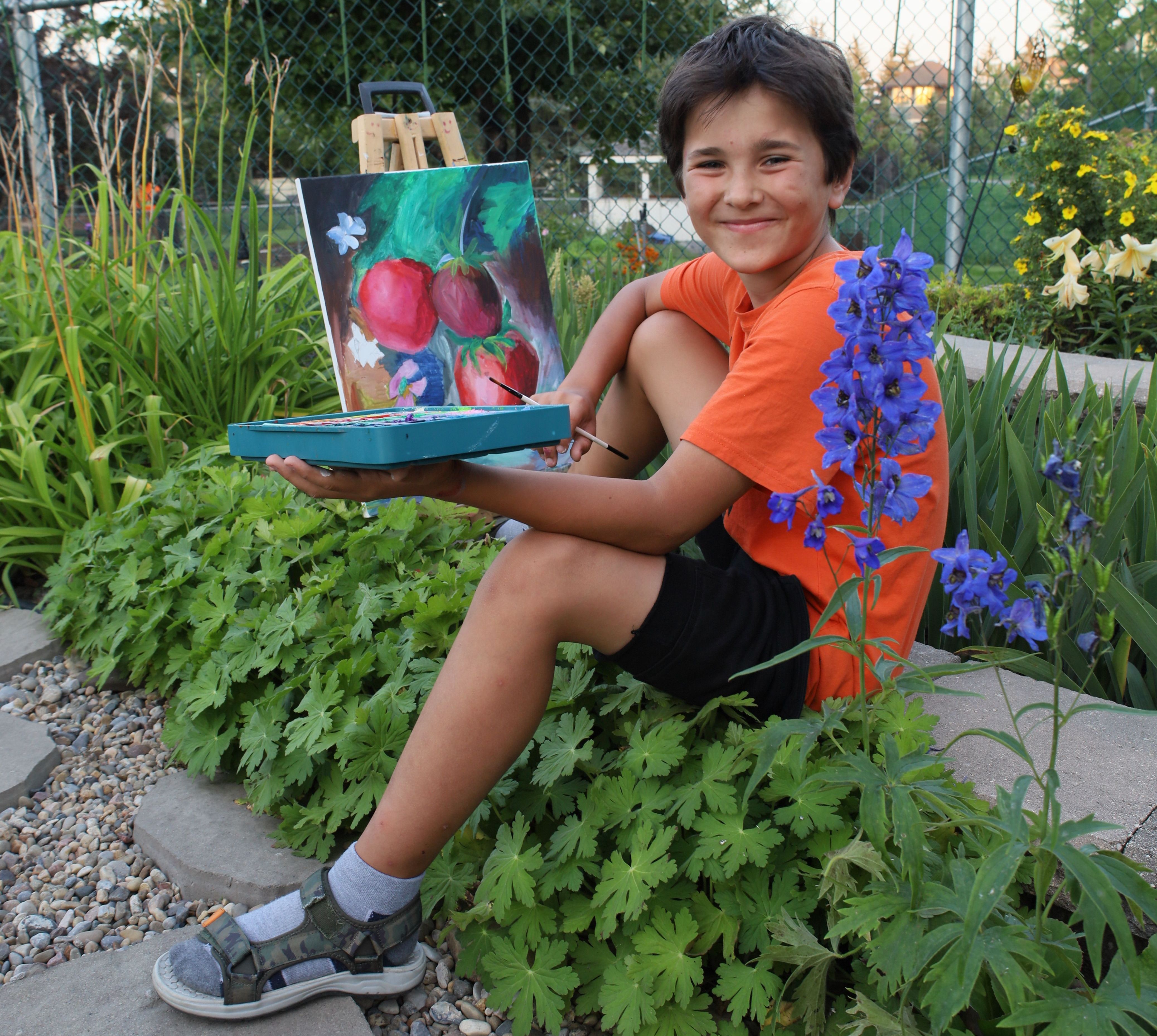 A smiling boy holding paints and a brush sitting among flowers