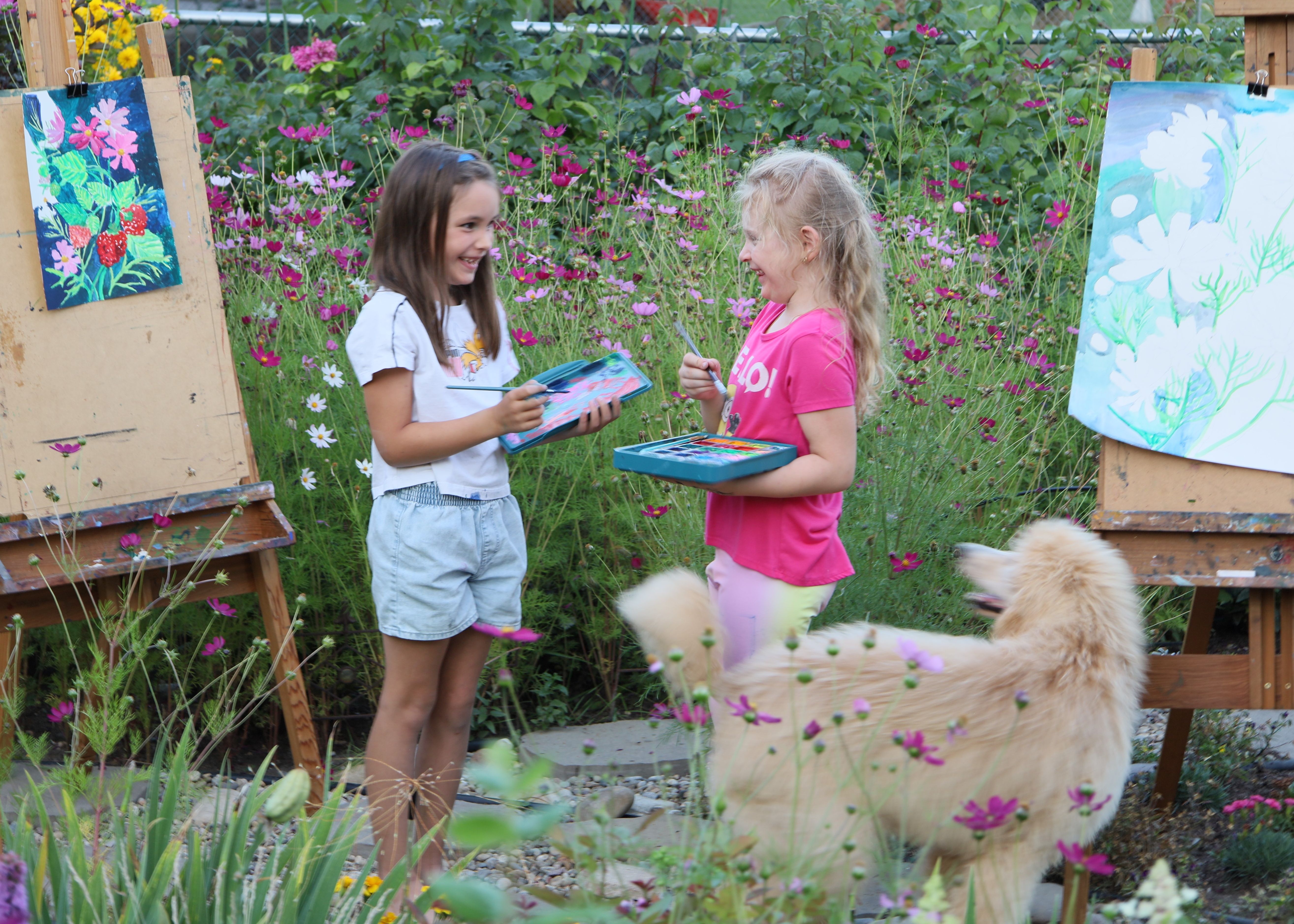Two girls holding brushes and paints standing in the garden, looking at each other and smiling
